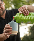 Person pouring liquid from a green container into a small blue and beige cup outdoors.
