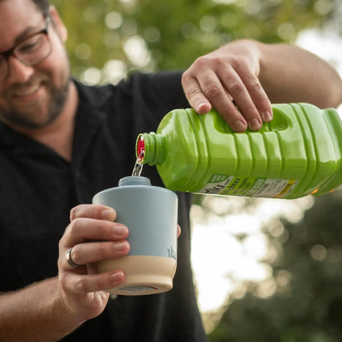 Person pouring liquid from a green container into a small blue and beige cup outdoors.