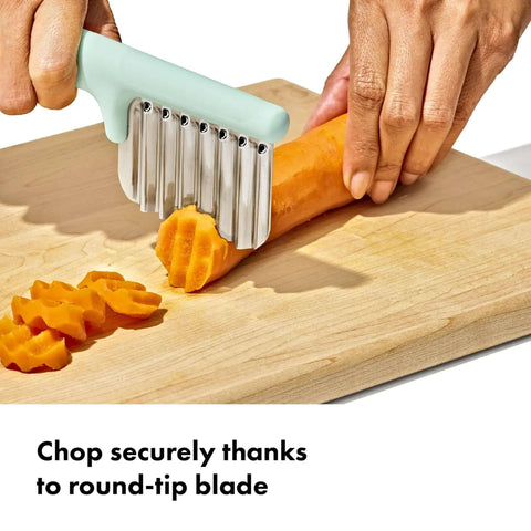 Oxo Tot crinkle cutter with mint-green handle and stainless steel blades slicing a carrot on a cutting board.