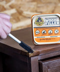 Front view of Gorilla Wood Filler 16 oz tub; hand applies wood filler to a wooden cabinet with a putty knife.