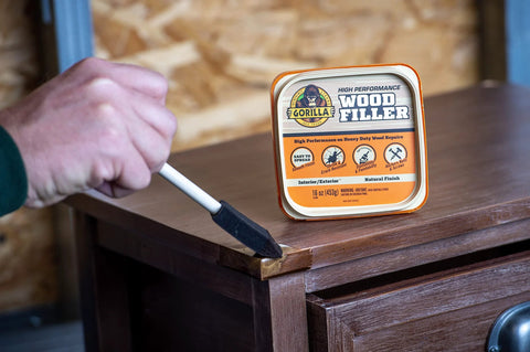 Front view of Gorilla Wood Filler 16 oz tub; hand applies wood filler to a wooden cabinet with a putty knife.
