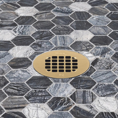 Universal 4 Inch Round Shower Strainer Snap In Bathroom Drain Cover Brushed Gold Stainless Steel installed on dark hexagonal tiles.