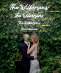 Couple under neon sign reading 'The Wilkersons' with various sizes shown, surrounded by greenery. Wedding or event setup indoors.