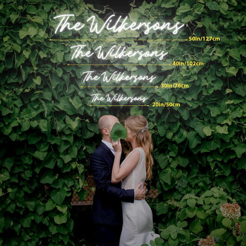 Couple under neon sign reading 'The Wilkersons' with various sizes shown, surrounded by greenery. Wedding or event setup indoors.