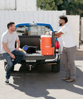 Two men with 10 Gallon Portable Sports Cooler And Beverage Dispenser With Flat Seat Lid on truck bed, orange and white cooler visible.