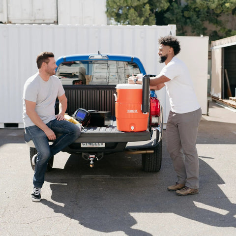 Two men with 10 Gallon Portable Sports Cooler And Beverage Dispenser With Flat Seat Lid on truck bed, orange and white cooler visible.