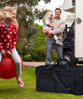 65L Duffle Bag With Shoes Compartment, black foldable travel bag on grass, campervan and family in background, water repellent.