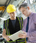 Two men reviewing plans in a safety environment; one wears Top Shelf Seven Pocket Mesh Safety Vest In Matte Black for Active Crews.