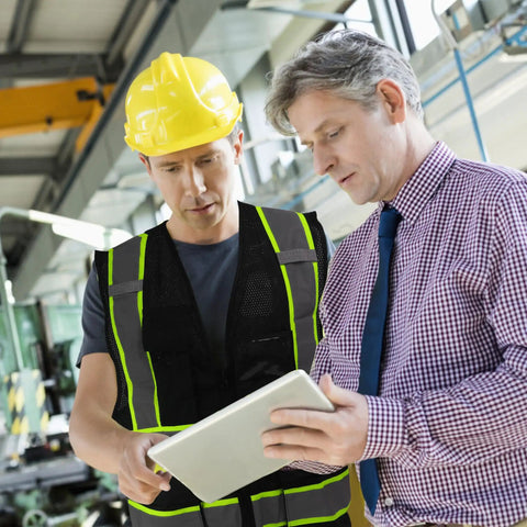 Two men reviewing plans in a safety environment; one wears Top Shelf Seven Pocket Mesh Safety Vest In Matte Black for Active Crews.