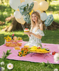 Woman enjoying outdoor picnic on a red blanket with fruit, cake, and balloons, surrounded by greenery in a park setting.