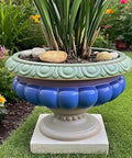 Large decorative garden planter with blue and green ornate design on a pedestal, filled with greenery, placed on grass.