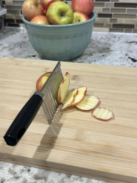Side view of stainless steel crinkle-cut blade slicer with black handle slicing an apple on a wooden cutting board.
