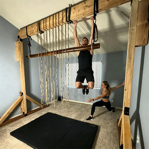 Man hanging from a wooden exercise frame with ropes and woman assisting indoors, on a padded mat in a home gym.