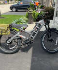 Dual Motor Electric Bike For Adults With Four Thousand Watt Power And Forty Six Mph Speed Featuring A Massive Fifty Two Volt Seventy Ah Battery That Crushes Steep Hills Delivering An Insane Two Hundred Mile Range Full Suspension Fat Tire Ebike parked outside with a green bag attached.