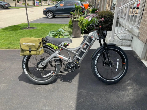Dual Motor Electric Bike For Adults With Four Thousand Watt Power And Forty Six Mph Speed Featuring A Massive Fifty Two Volt Seventy Ah Battery That Crushes Steep Hills Delivering An Insane Two Hundred Mile Range Full Suspension Fat Tire Ebike parked outside with a green bag attached.