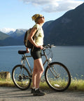 Person with bicycle by a lake, wearing outdoor gear and helmet, with mountains in the background.
