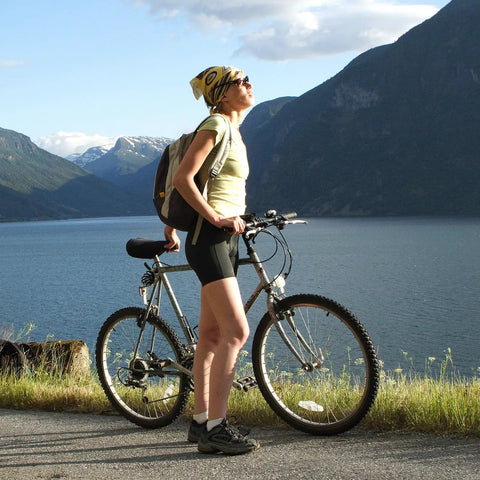 Person with bicycle by a lake, wearing outdoor gear and helmet, with mountains in the background.