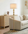 Riley 60 Inch Floor Lamp With Drum Shade In Gold Brass Finish For Living Room Or Bedroom Modern Standing Lamp With Boho Chic Mid Century Vibe And Elegant Corner Light For Office next to a beige sofa.
