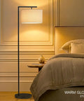 Modern LED Floor Lamp With Tan Fabric Shade Tall Corner Pole Standing Light For Living Rooms And Offices Ideal Bedroom Reading Light Black Finish next to bed with warm glow.