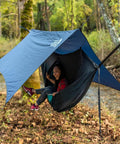 Person relaxing in All-In-One OneLink Hammock Shelter System With DoubleNest Hammock Atlas Suspension Straps Guardian Bug Net ProFly Rain Tarp And Stakes Packable Lightweight Gear For Easy Setup Camping Backpacking And Outdoor Adventures by a river.