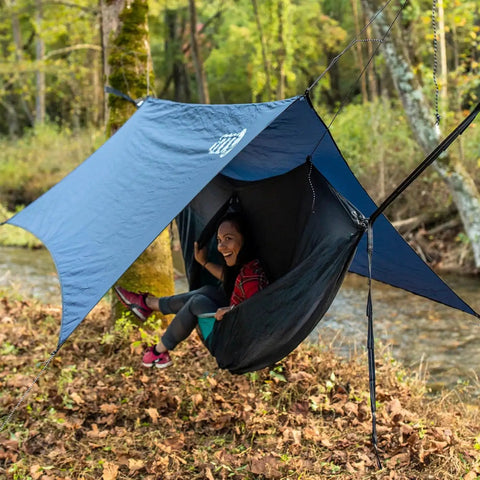 Person relaxing in All-In-One OneLink Hammock Shelter System With DoubleNest Hammock Atlas Suspension Straps Guardian Bug Net ProFly Rain Tarp And Stakes Packable Lightweight Gear For Easy Setup Camping Backpacking And Outdoor Adventures by a river.