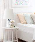 Totally Sparkly Pink Unicorn Table Lamp With Glitter Resin Base And White Tapered Shade on nightstand beside bed.