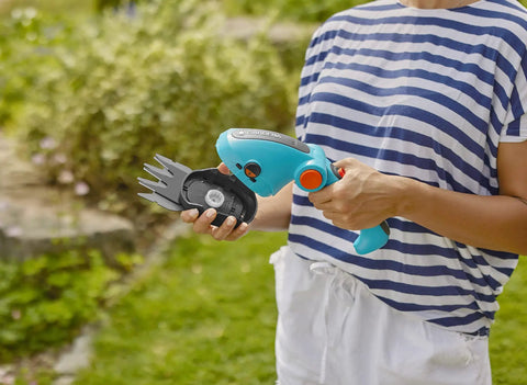 Person holding a cordless hedge trimmer and grass shears, battery powered handheld electric garden tool with ergonomic design.