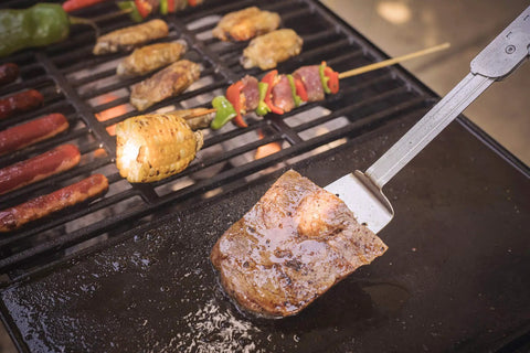 Six In One Folding Grill Utensils Set Featuring Fork Spatula Knife Tongs Bottle Opener And Corkscrew For Fourteen Inch Portable Backyard BBQ And Camping Gear next to grilled meats and vegetables
