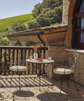 Outdoor patio with two wrought iron chairs and a table, overlooking lush green hills and a stone building with wooden windows.