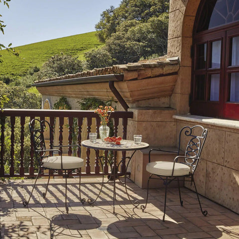 Outdoor patio with two wrought iron chairs and a table, overlooking lush green hills and a stone building with wooden windows.