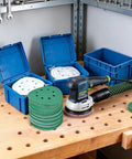 Rock Solid 5 Inch 8 Hole Zirconia Alumina Sanding Discs assorted grits beside random orbital sander on workbench.