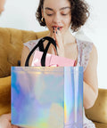 Woman holding and looking at a Poppin Glossy Iridescent Gift Bags Set Of 15 Reusable Goodie Bags With Handles, 12.6x4.7x11 inches.