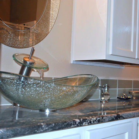 Clear Glass Oval Slipper Vessel Sink Set With Slick Brushed Nickel Hardware Tempered Glass Scratch Resistant Finish One Point Five Inch Brass Pop Up Drain Includes Mounting Ring Perfect Modern Vanity Centerpiece on granite countertop.