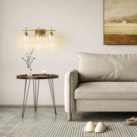 Three Light Vanity Light With Clear Glass Shades In Modern Gold Finish For Bathroom Over Mirror Contemporary Wall Lamp For Soft Balanced Lighting In Bathroom Bedroom Or Hallway mounted on wall beside sofa and table.