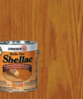 No Sweat Shellac Sealer And Heavy Duty Odor Blocker For Wood Trim And Paneling Covers Hundreds Of Square Feet Fast Drying Non Toxic Formula For A Smooth Mid Tone Sheen In Badass Amber One Gallon in a gallon can on a wooden background.