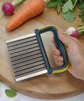 Crinkle cutter knife with stainless steel ridged blade and blue-yellow handle held over a wooden cutting board with carrots and radishes