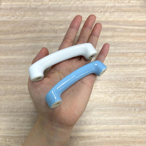 Hand holding two ceramic cabinet pulls, white glossy and light blue, showcasing design and size contrast.