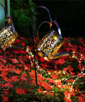 Two copper-toned solar waterfall watering can lights glowing over red flowers at night. 2 Pack Solar Waterfall Watering Can Lights For Garden, LED Copper Outdoor Stake Lamps, Retro Waterproof Yard Decor For Pathway Lawn Patio Deck Walkway And Courtyard Dope Garden Accent Lighting.