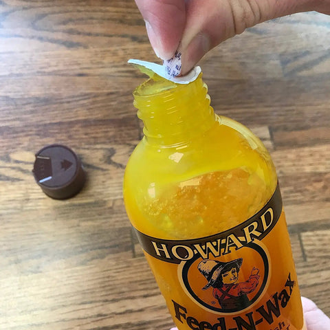 Person peeling seal off orange scent wood polish and conditioner cream formula with natural oils, bottle on wood floor.