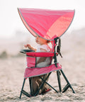 Child sitting in a pink Go With Me Venture Portable Toddler Chair With Sun Canopy on the beach, foldable and versatile for outdoor use.