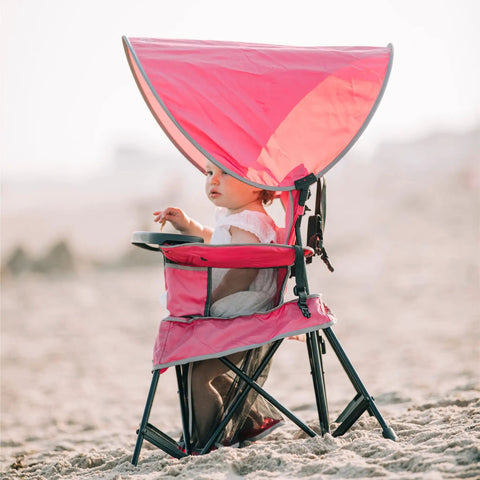 Child sitting in a pink Go With Me Venture Portable Toddler Chair With Sun Canopy on the beach, foldable and versatile for outdoor use.
