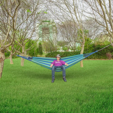 Person relaxing on Extra Long And Wide Double Hammock For Travel Camping Backyard Porch Or Indoor Use With Carrying Pouch Included Blue Green Stripes Ultra Comfy Cotton Weave 142 By 63 Inches Overall And 94 By 63 Inch Bed strung between trees