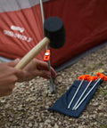 Hammer driving Beefy 10-Inch Steel Tent Stakes Heavy-Duty Ground Anchors next to tent on rocky ground.