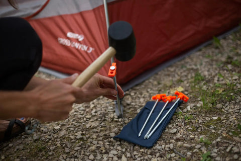 Hammer driving Beefy 10-Inch Steel Tent Stakes Heavy-Duty Ground Anchors next to tent on rocky ground.