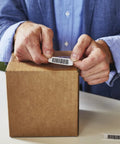 No Sweat Multi Purpose Thermal Labels For Labelwriter Printers 1 By 2 1 8 Inch being applied to a shipping box by hands.