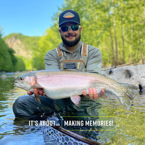 Angler holding a large caught fish in a stream surrounded by green foliage.