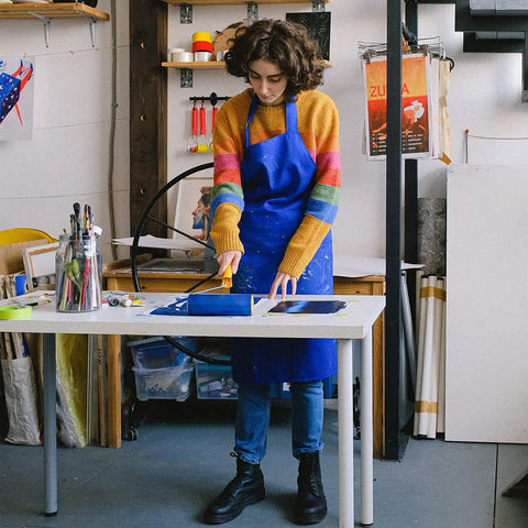 Person using Blue Multi-Surface Painter's Tape 0.94 In X 60 Yds, 6 Rolls, in art studio, wearing blue apron and colorful sweater