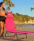 Woman setting up pink 2-leg chaise lounge chair with polyester comfort, reclining back, headrest pillow on beach