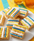 Squares of rice crispy treats with blue, yellow, and white icing stripes on a white plate with colorful background.