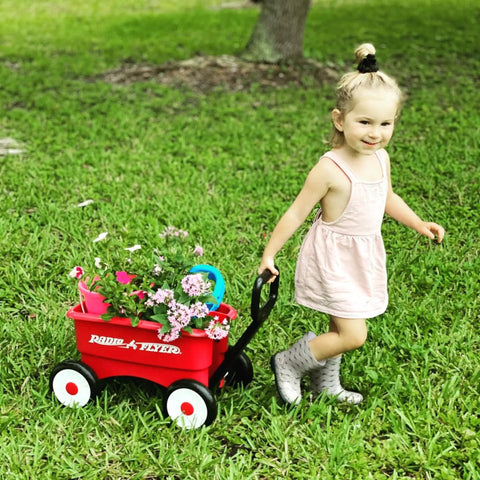Child pulling a red push and pull walker wagon with garden tools and flowers, suitable for kids aged one to four years old.